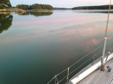 Very still water reflecting pink clouds, with shorelines mirrored in the water. The deck of a sailboat is visible in the lower right corner.