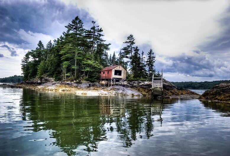 Old boathouse with open doors on a small wooded point of land, with placid water reflecting the trees on shore.