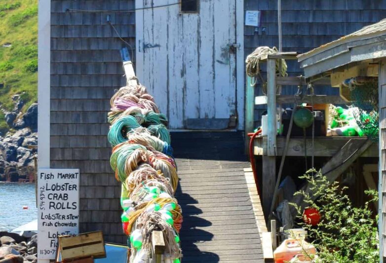 Weathered wooden building with closed white door. A ramp leads up to the door and the railing is covered with colorful lobster buoys and ropes..
