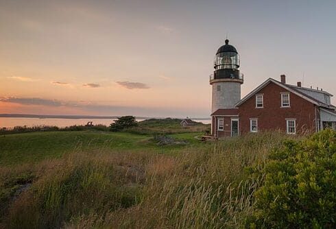 Lighthouse on Seguin Island is a brick house with white tower. Grassy foreground with sunset glowing pink over the water.