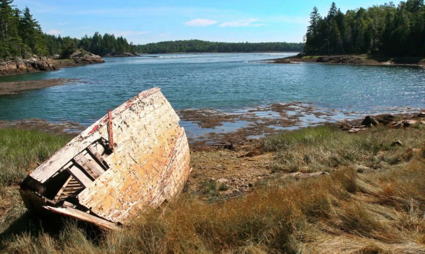 The ruins of a wooden boat rests on the shore of a scenic harbor.