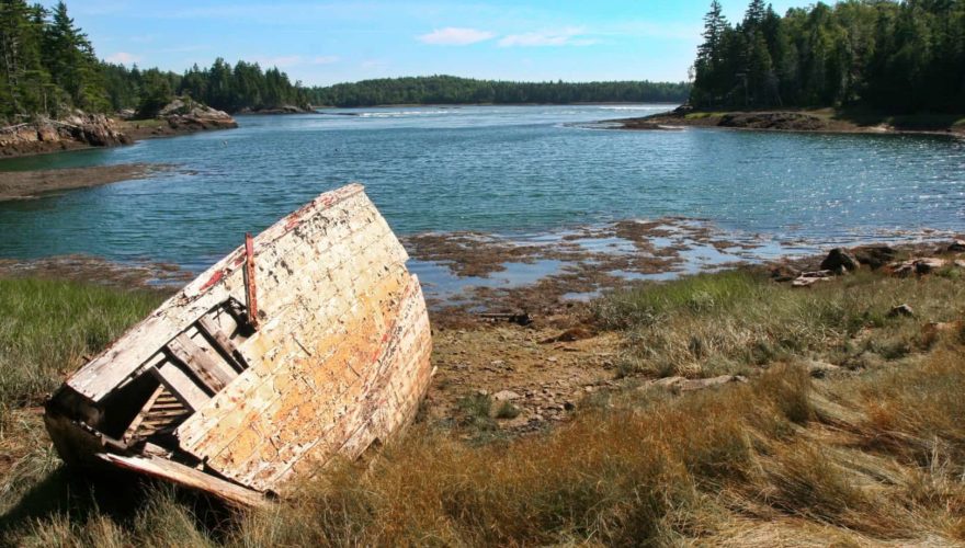 The ruins of a wooden boat rests on the shore of a scenic harbor.