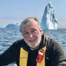 man with white beard wearing safety harness with icebergs behind him