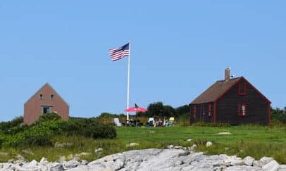 Smuttynose at Isles of Shoals with a rocky shore, grassy lawn, and two building flanking a flagpole flying an American flag.