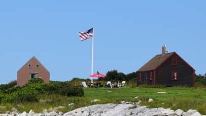 Smuttynose at Isles of Shoals with a rocky shore, grassy lawn, and two building flanking a flagpole flying an American flag.