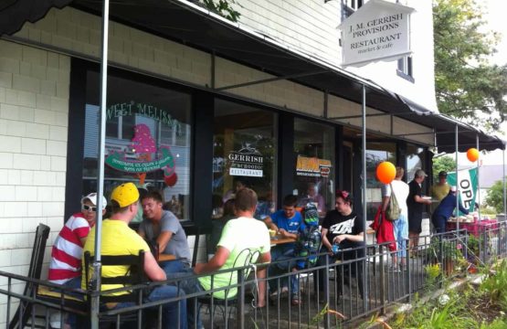 Covered porch crowded with people at tables outside store entrance.