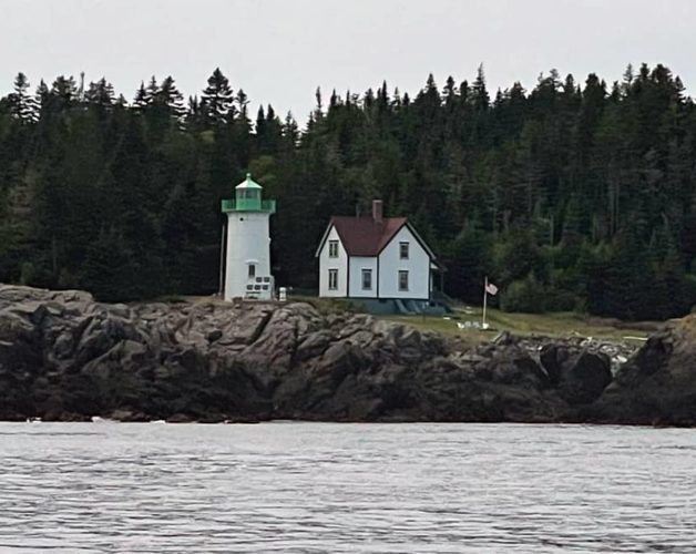 Classic lighthouse tower and house on a rocky shore with a pine forest behind.