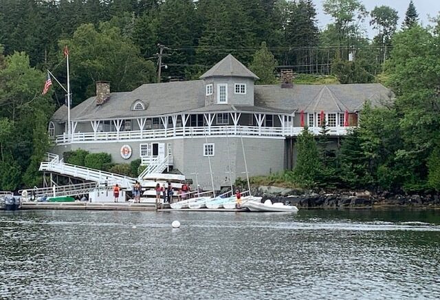 Winter Harbor Yacht Club building is a large shingle-style structure perched on the water's edge. Docks with sailing dinghies are below it.