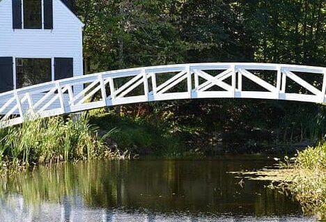 The iconic arched white bridge over quiet water in Somesville, with white building to its left side.